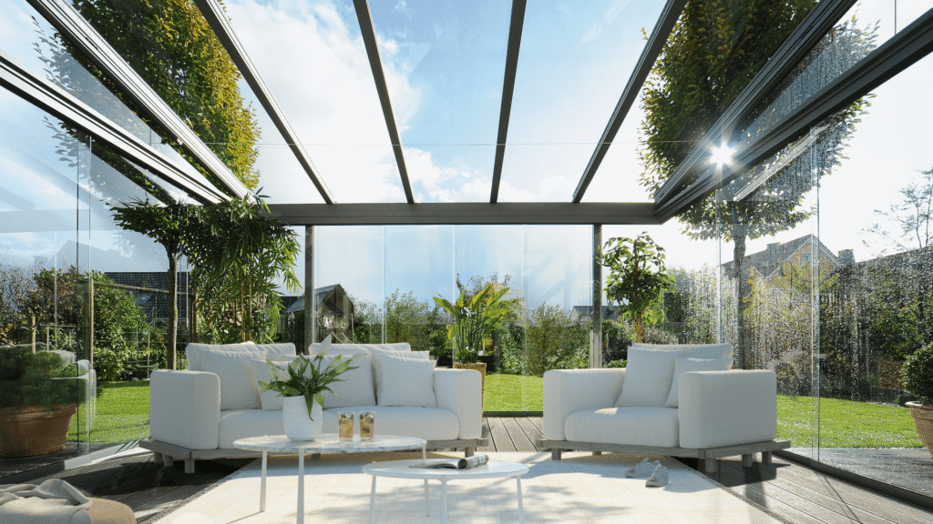 Teras Kapatma | Bursa 4 Mevsim Teras Çözümleri (Balkon Art) 2 Cam tavanlı ve cam duvarlı modern kış bahçesi, içerisinde oturma grubu ve dışarıda yeşil bahçe manzarası.